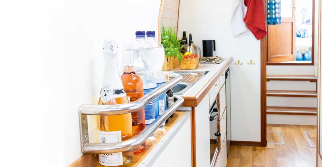 Die Küchenzeile einer Kormoran mir Spüle, Backofen, Gasherd und Kühlschrank. Im Vordergrund stehen einige Flaschen in einem Flaschenhalter