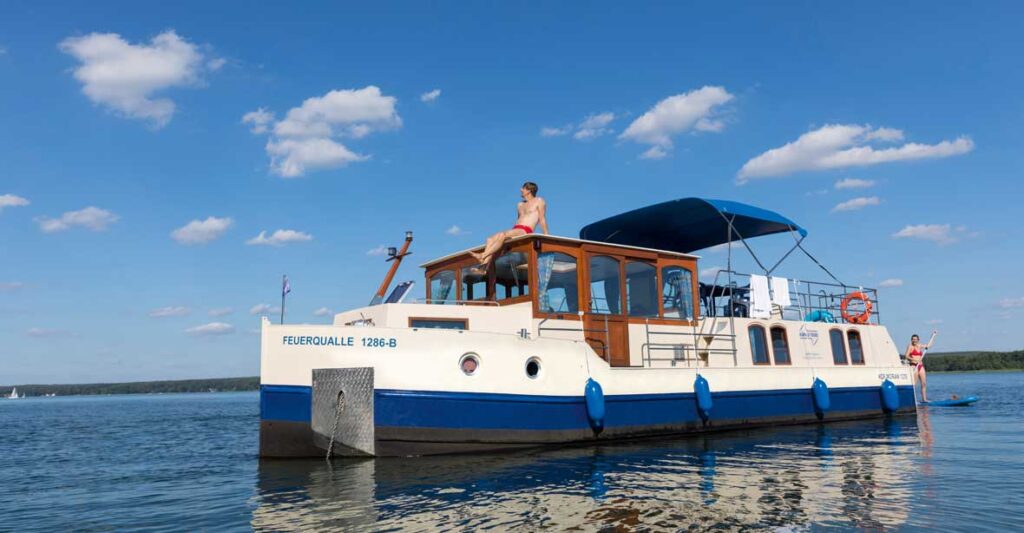 Eine Seitenansicht der Kormoran 1290. auf dem Dach der Kormoran sitzt ein junger Mann, hinter der Kormoran kommt eine junge Frau auf einem SUP hervor.